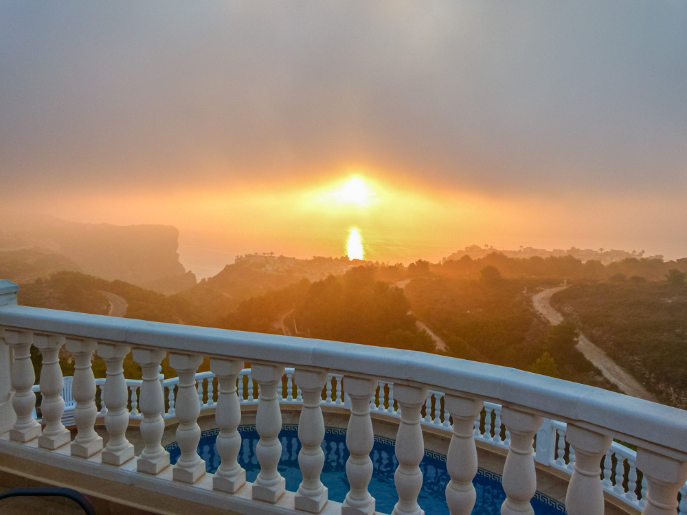 Blick von der Terrasse mit weißer Balustrade auf den Pool und den stimmungsvollen Sonnenuntergang über der Mittelmeerküste.