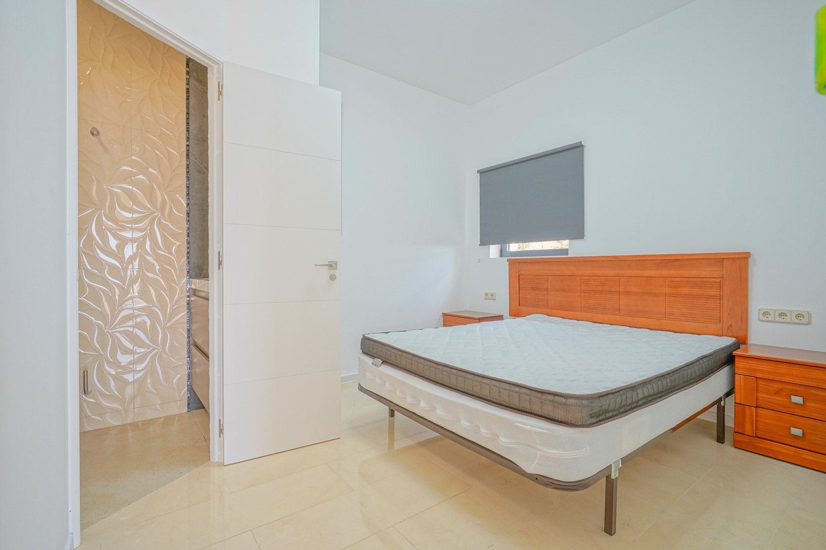 Doppelzimmer mit Holzmöbeln, hellen Fliesenböden und angrenzendem Badezimmer mit dekorativen Wandfliesen in Wellenoptik.