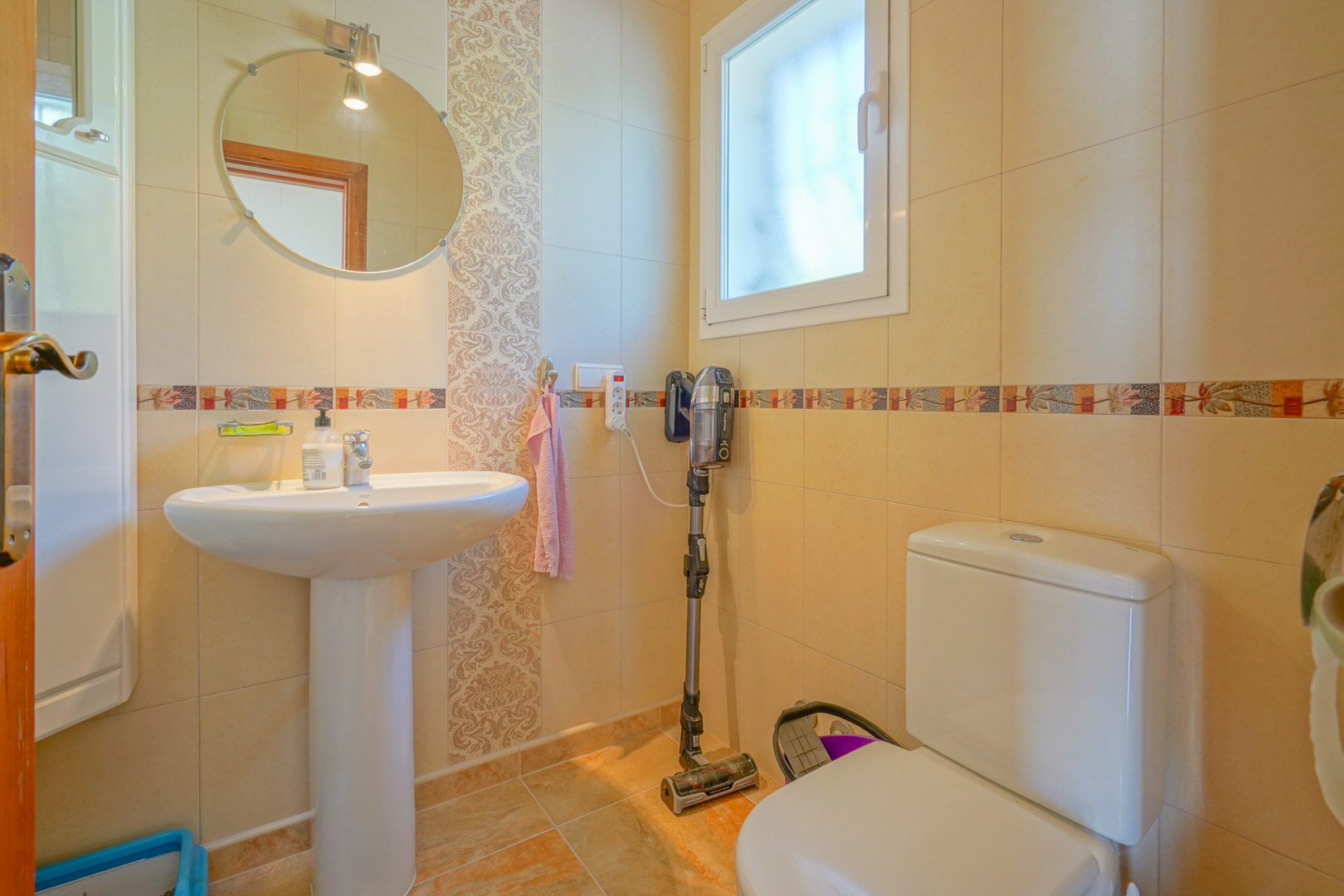 Funktionales Badezimmer mit Säulenwaschbecken, Rundspiegel und cremefarbenen Fliesenwänden mit dekorativen Bordüren und Fenster.