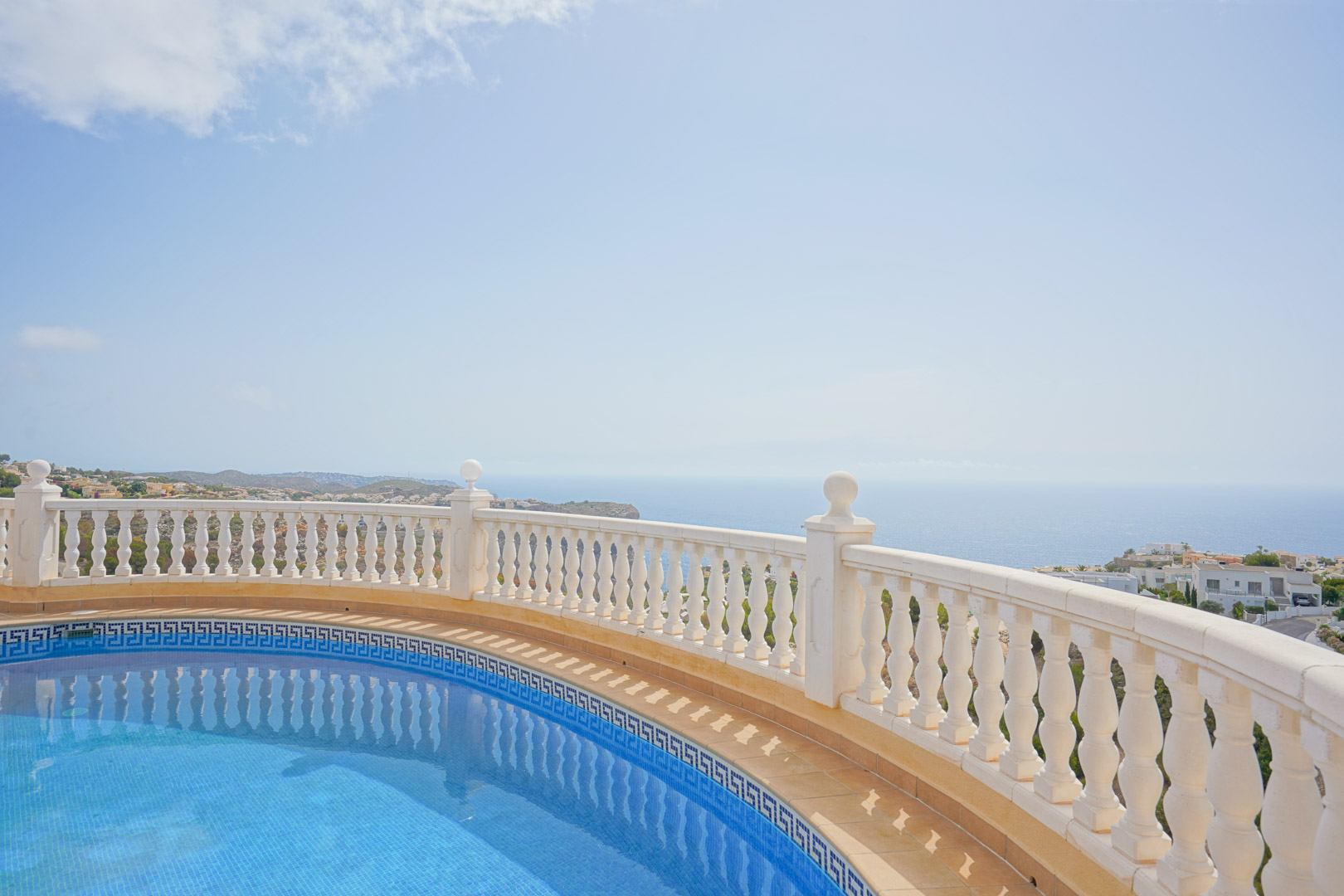 Geschwungener Pool mit blauen Mosaikfliesen und Mäander-Bordüre, begrenzt durch eine weiße Balustrade mit Panoramablick auf das Mittelmeer.