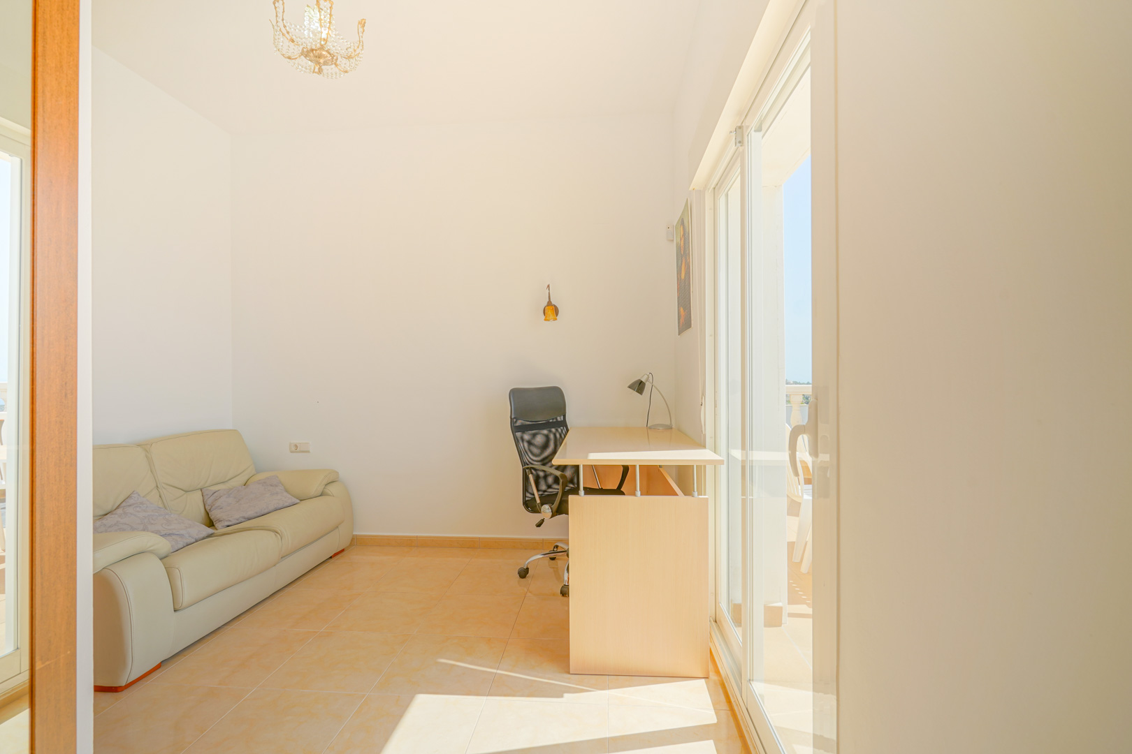 Heller Mehrzweckraum mit Arbeitsbereich, Ledersofa und direktem Terrassenzugang durch große Glasschiebetüren. Warme Keramikfliesen unterstreichen das natürliche Tageslicht.