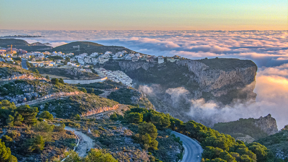 Panoramablick auf die Klippen von Cumbre del Sol über einem Wolkenmeer bei Sonnenuntergang, der die zerklüftete Küstenlinie und die Wohngebiete zeigt.