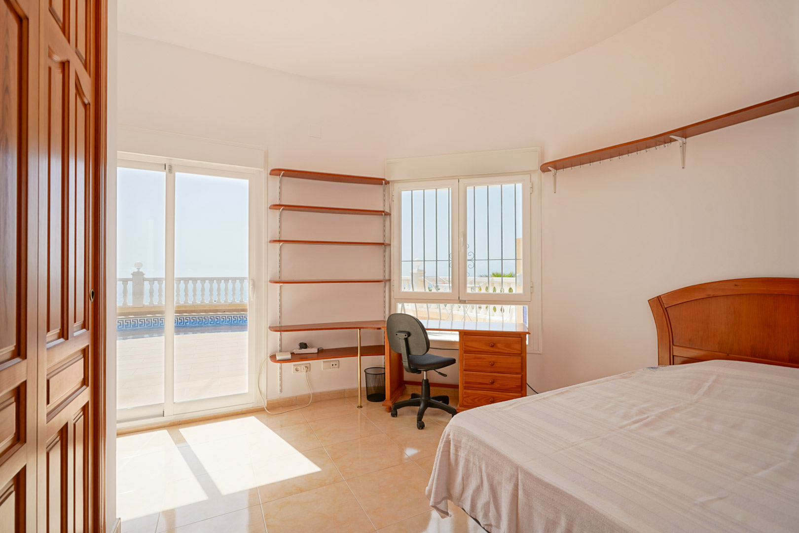 Schlafzimmer mit Arbeitsbereich, Holzregalen und direktem Terrassenzugang durch Glasschiebetüren. Helle Keramikböden und viel Tageslicht.