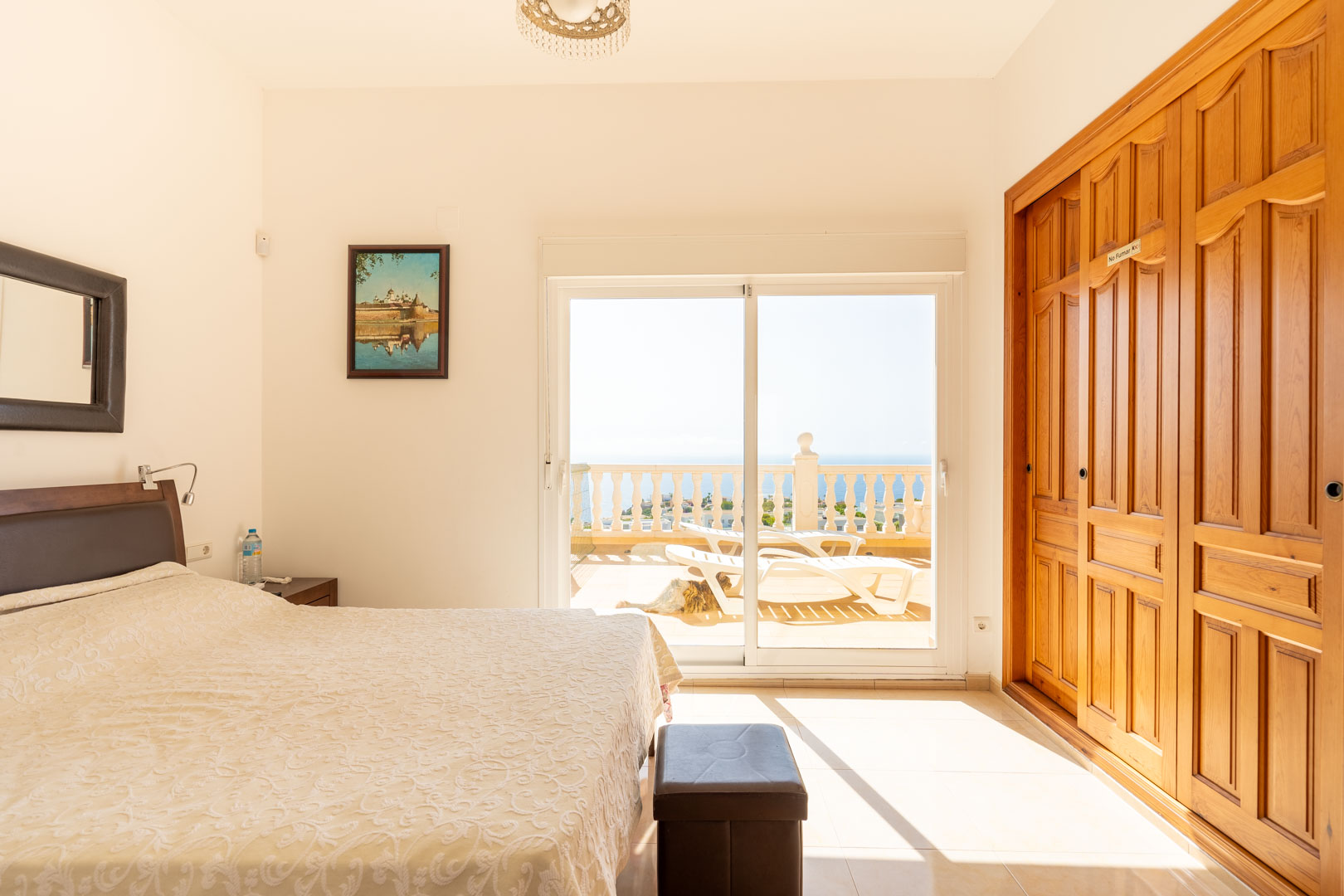 Schlafzimmer mit Einbauschränken aus Holz und Glasschiebetüren, die auf eine sonnige Terrasse mit Meerblick und Liegestühlen führen.