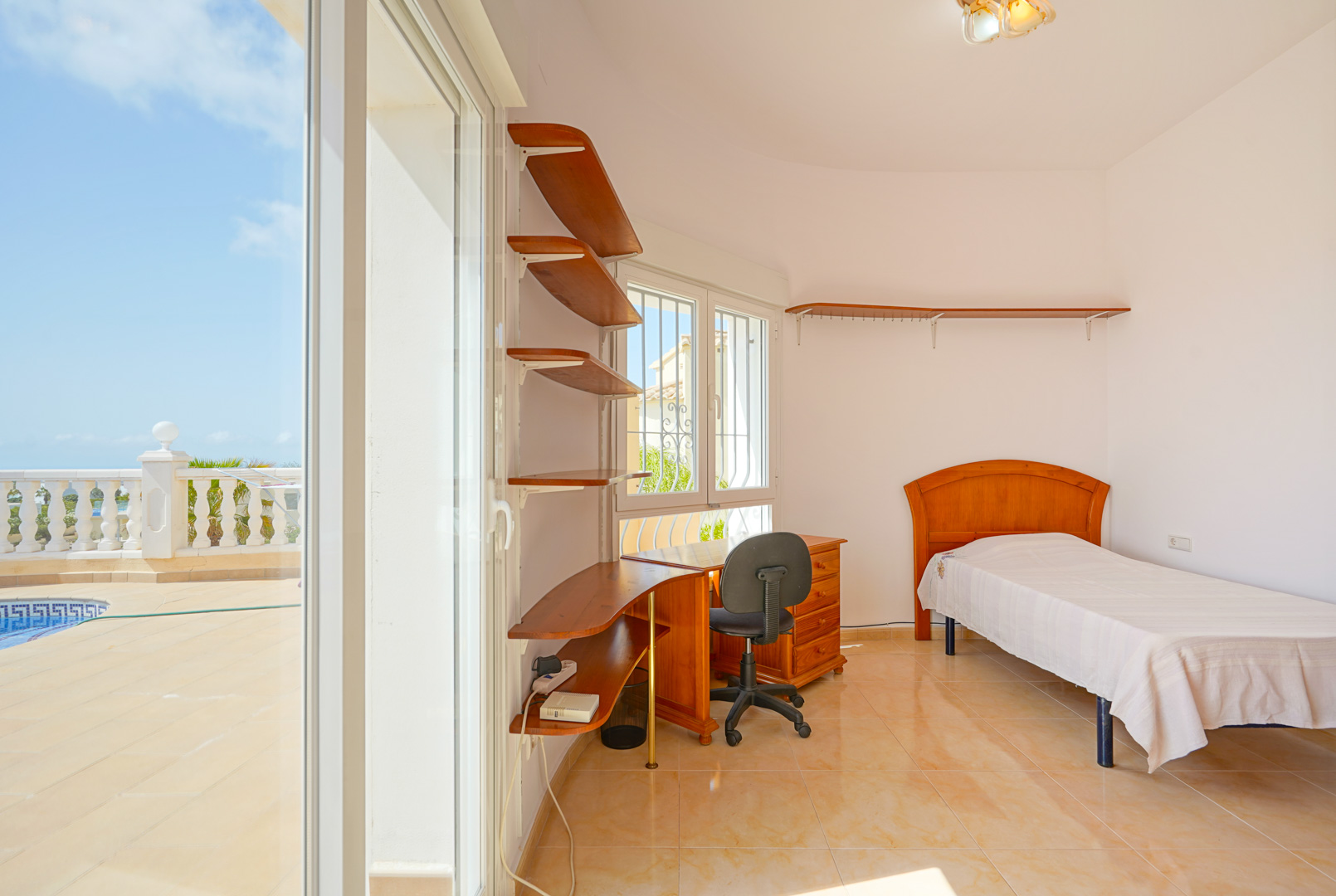 Schlafzimmer mit Einzelbett, Holzschreibtisch und Wandregalen. Große Glasschiebetüren ermöglichen den direkten Zugang zur Poolterrasse.