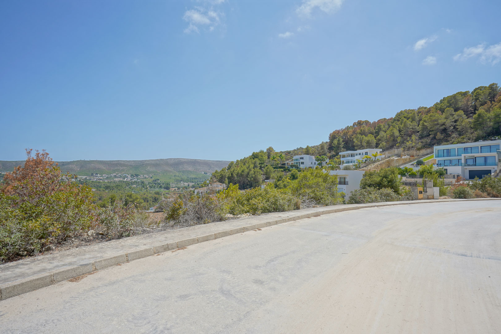 Blick von der asphaltierten Zufahrtsstraße der Erschließung auf das steile Gelände und die modernen Villen mit Panoramablick über das Tal und die Hügel.