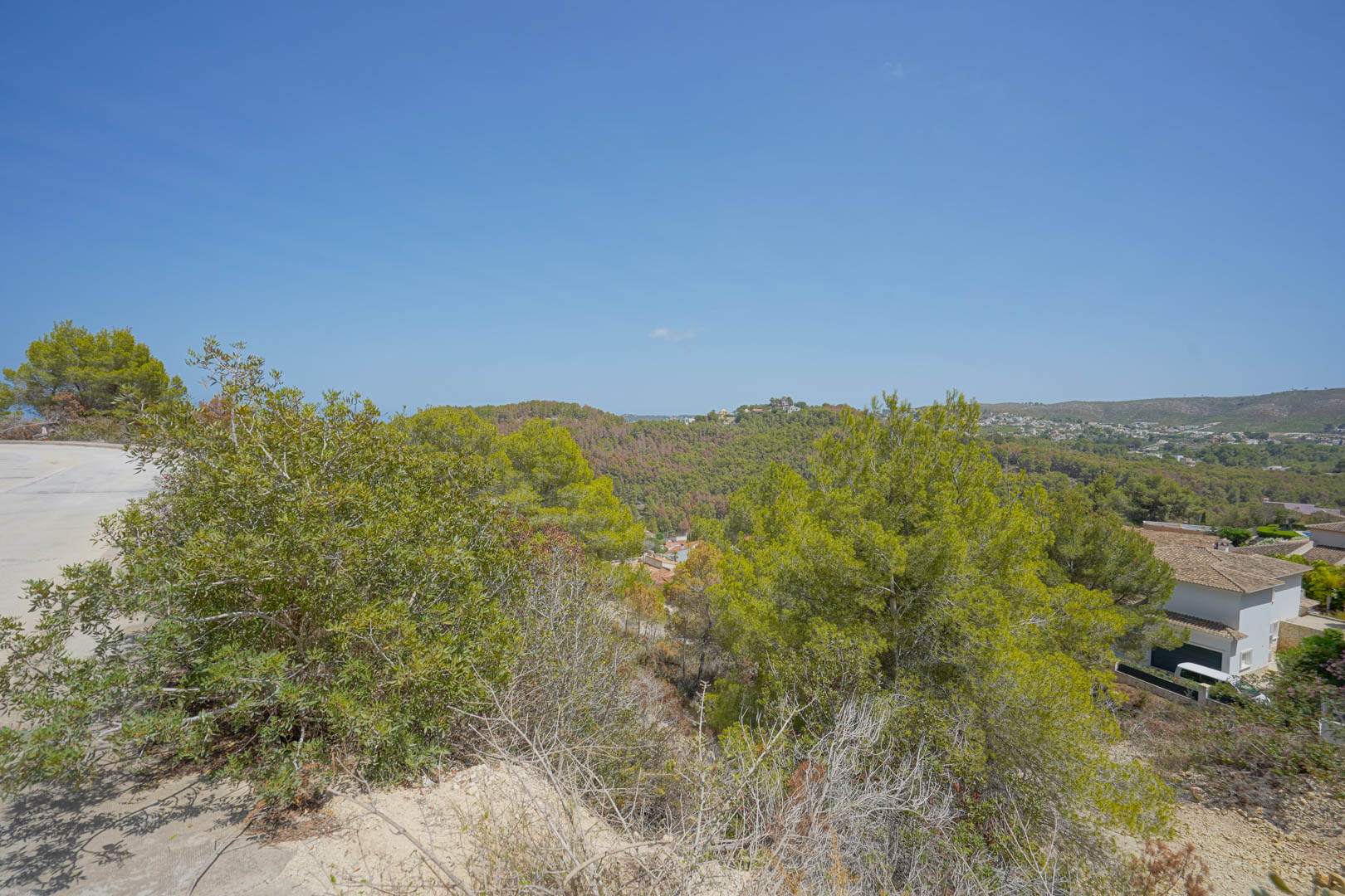 Erhöhte Aussicht vom Grundstück mit asphaltiertem Straßenzugang und Panoramablick auf die umliegenden grünen Hügel und mediterrane Flora.