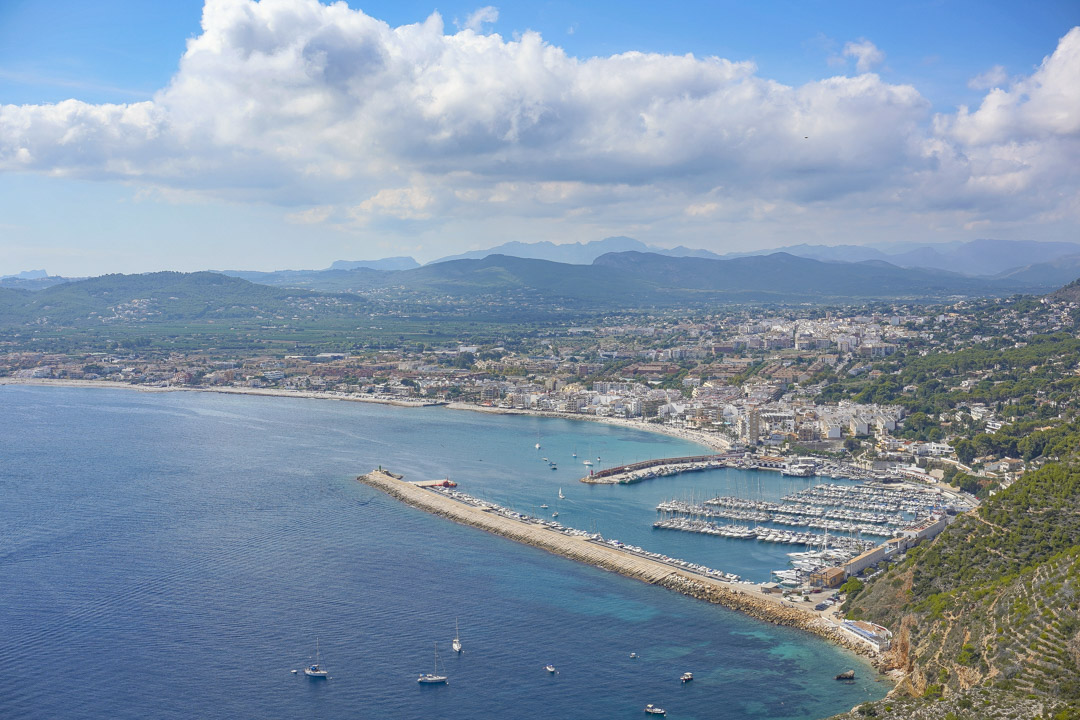 Erhöhter Panoramablick auf den Yachthafen und die Küstenlinie mit der langen Hafenmauer, zahlreichen Booten und den umliegenden Bergen im Hintergrund.