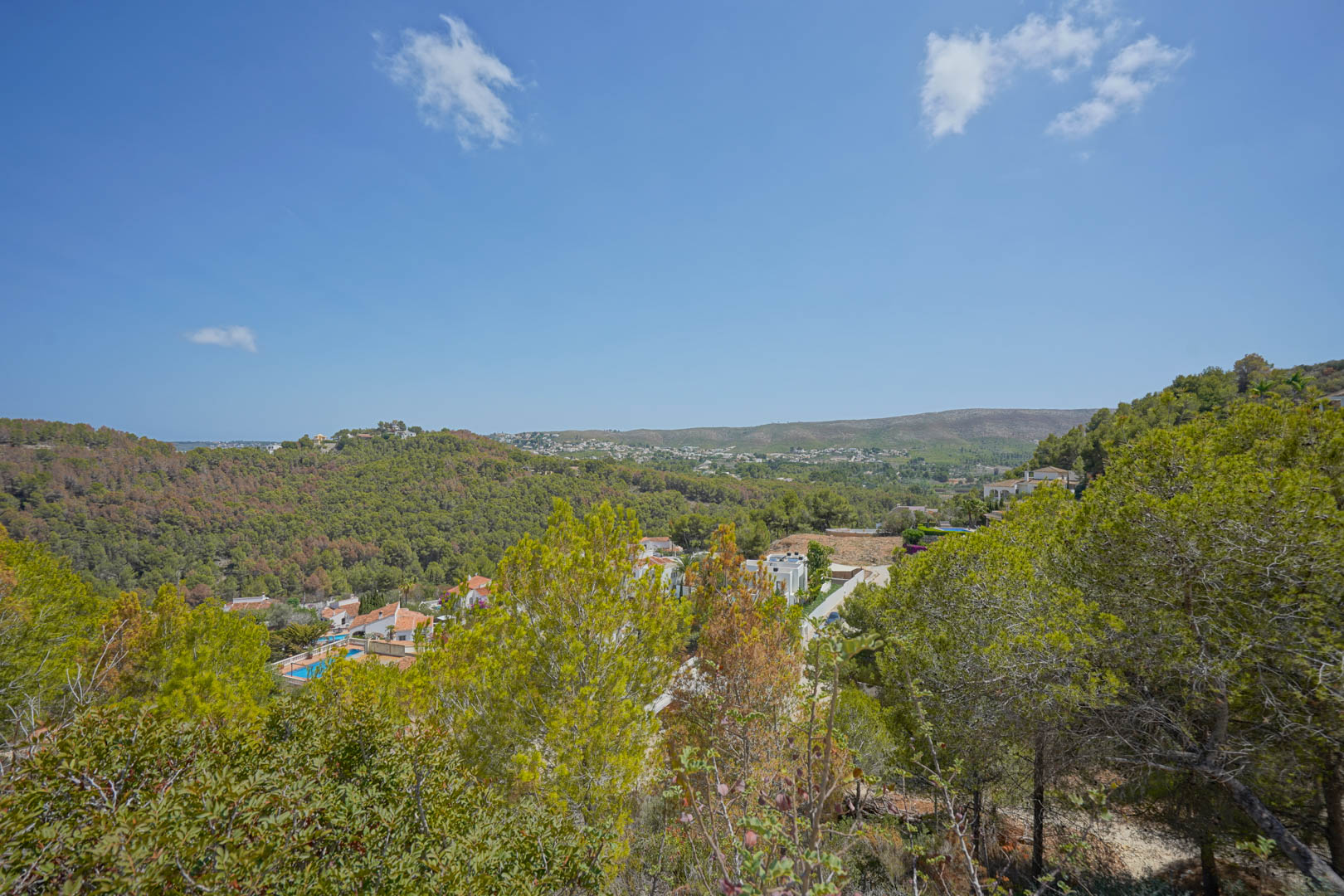 Erhöhter Panoramablick über ein grünes Tal mit mediterranen Pinien, weißen Villen mit Pools und sanften Hügelketten unter einem klaren, blauen Himmel.
