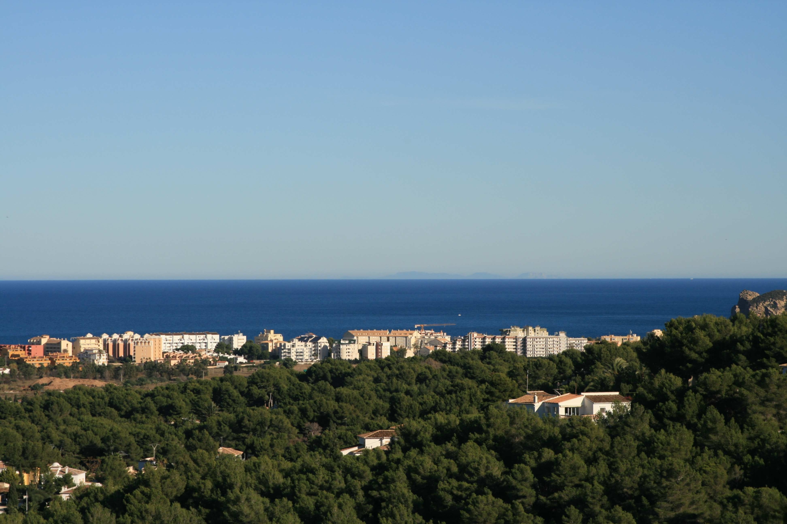 Panoramablick vom erhöhten Grundstück über einen dichten Pinienwald, das Küstenwohngebiet und das Mittelmeer am Horizont.