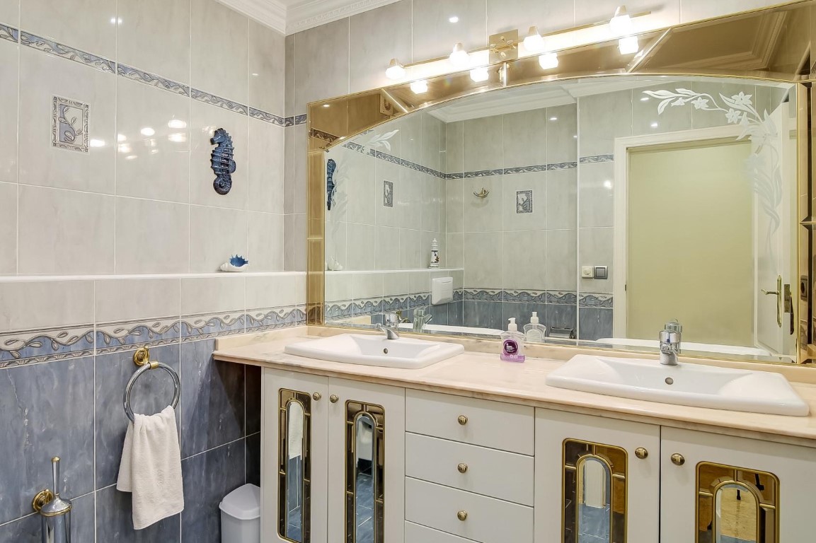 Hauptbadezimmer mit Doppelwaschbecken auf Marmorplatte, Schränken mit Goldakzenten, großem Spiegel und dekorativen blau-weißen Wandfliesen.