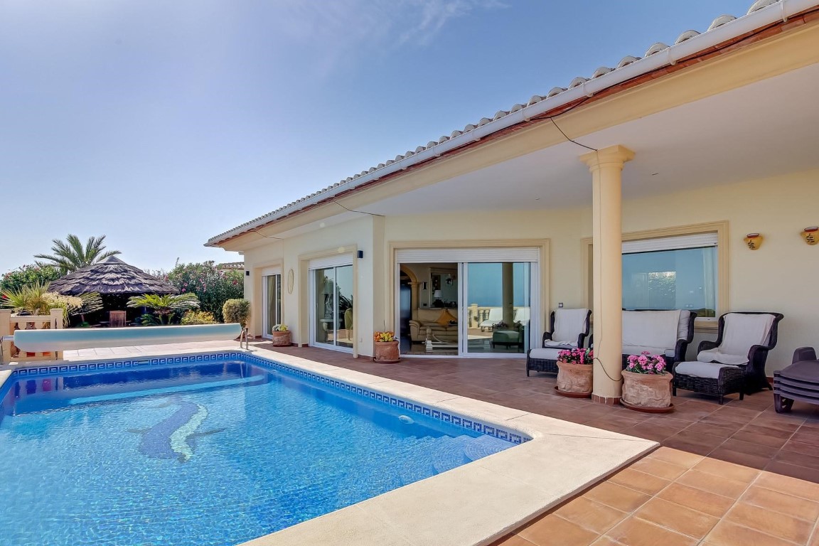 Hauptterrasse mit Mosaikpool, überdachter Veranda mit Säulen und Außenbereich. Im Hintergrund befindet sich ein Pavillon mit Reetdach inmitten mediterraner Bepflanzung.