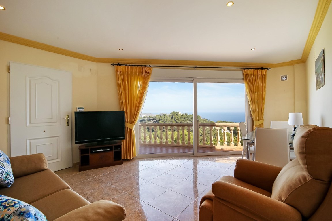 Heller Wohnbereich mit Fliesenboden, Zierleisten und großen Glasschiebetüren, die einen weiten Blick auf das Mittelmeer und die grüne Landschaft bieten.