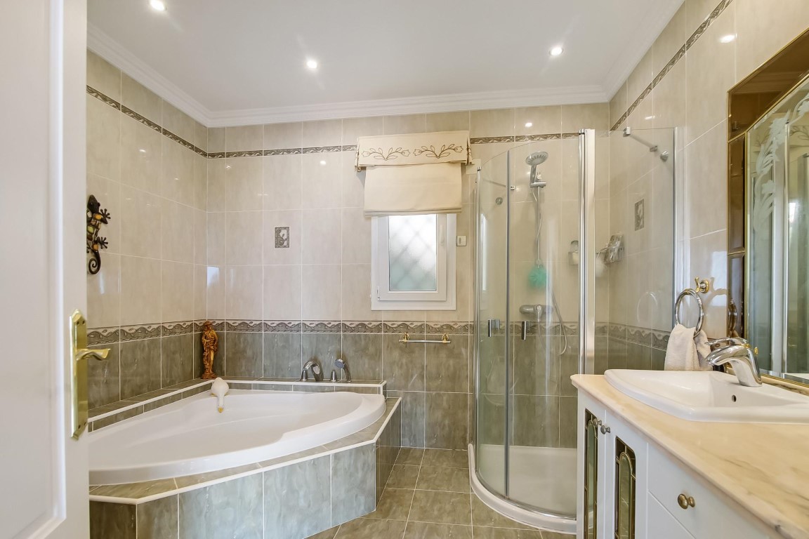 Komplettes Badezimmer mit Eckbadewanne, abgerundeter Duschkabine und Waschtisch mit Marmorplatte. Geflieste Wände mit Zierbordüren und Deckeneinbaustrahlern.