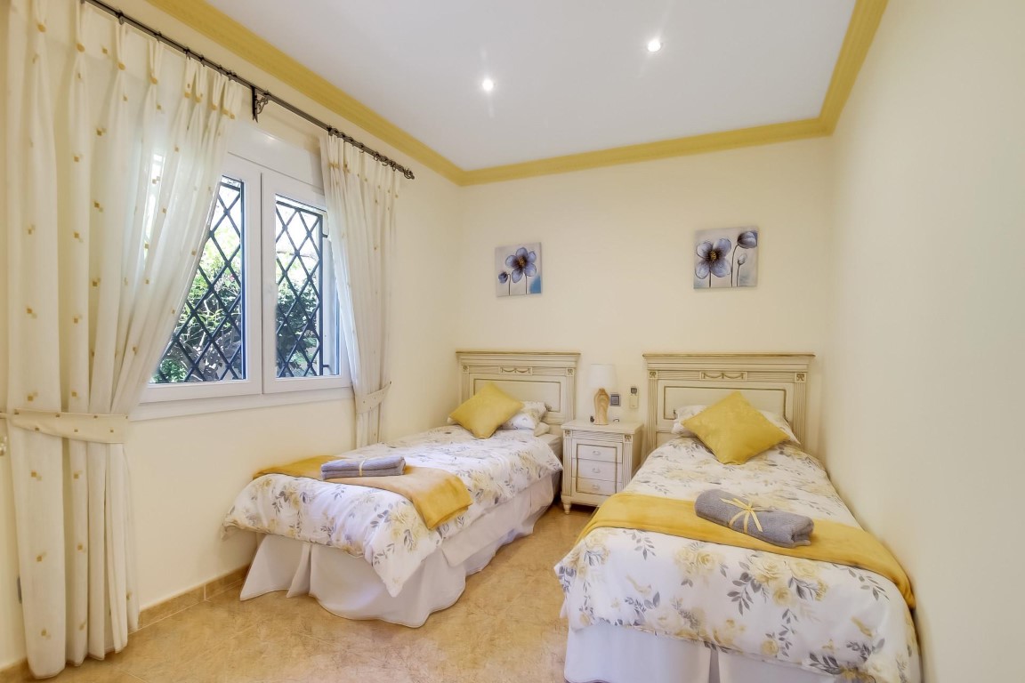 Schlafzimmer mit zwei Einzelbetten, weißen Kopfteilen, Deckenstrahlern und Fenster mit Sicherheitsgittern und hellen Vorhängen.