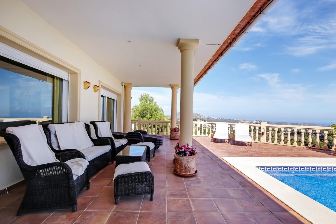 Überdachte Terrasse mit klassischen Säulen, Korbmöbeln und Terrakottaboden neben dem Pool, mit Panoramablick auf das Mittelmeer und die Landschaft.