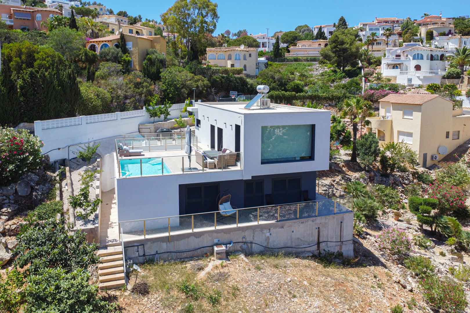 Außenansicht der modernen kubischen Villa mit Poolterrasse, Glasgeländern und Solarthermieanlage auf dem Flachdach in Hanglage.