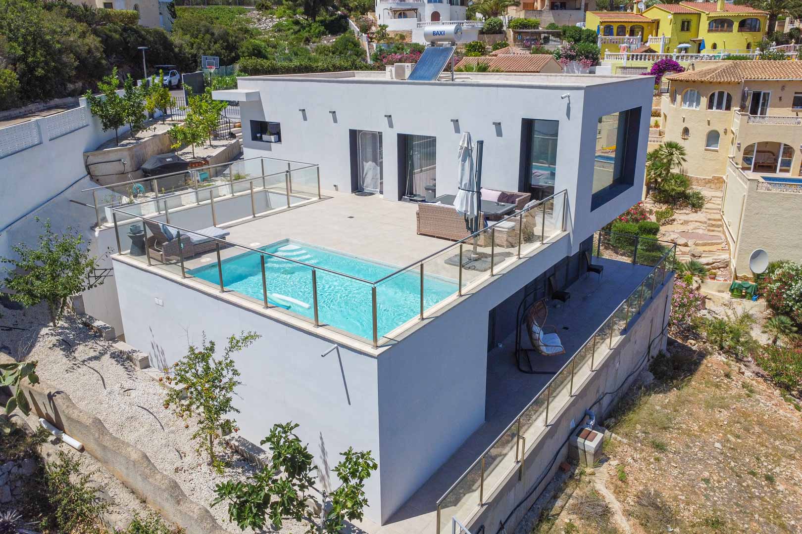 Außenansicht der modernen Villa mit Pool auf der oberen Terrasse, Glasgeländern, Solarpanel auf dem Dach und einer überdachten Terrasse im Erdgeschoss.