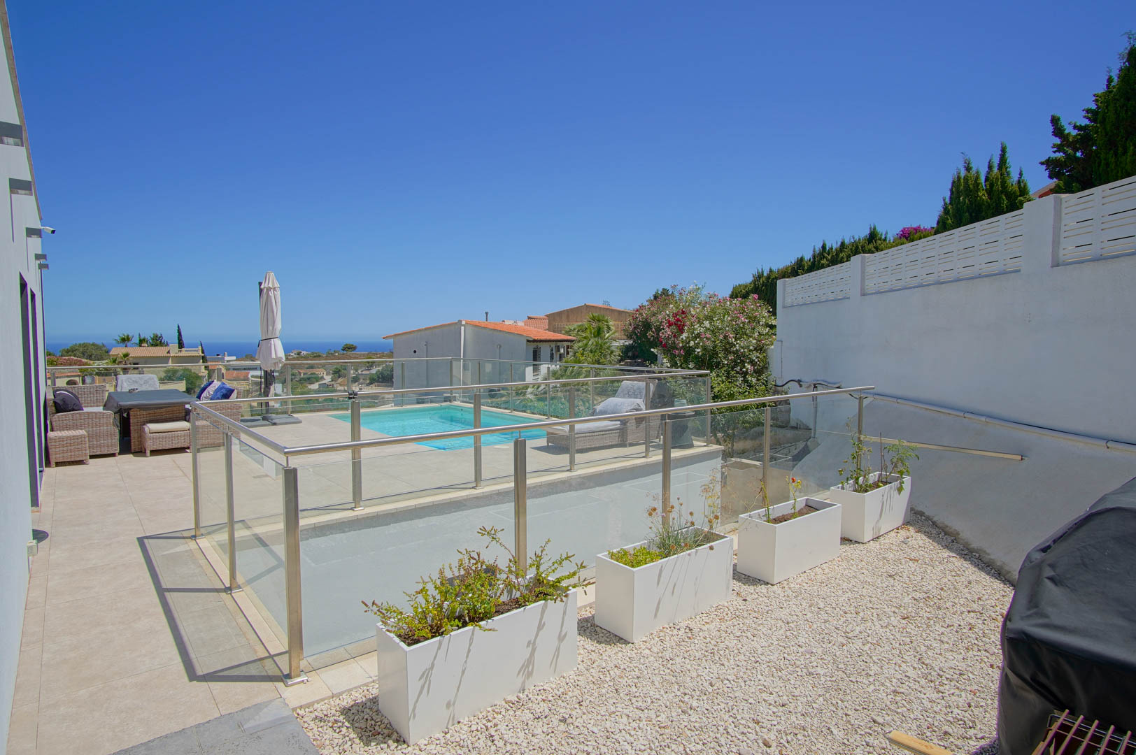 Außenbereich mit Glas- und Edelstahlgeländer, Poolterrasse mit Sonnenbereich und angrenzender Kiesfläche mit modernen weißen Pflanzgefäßen.