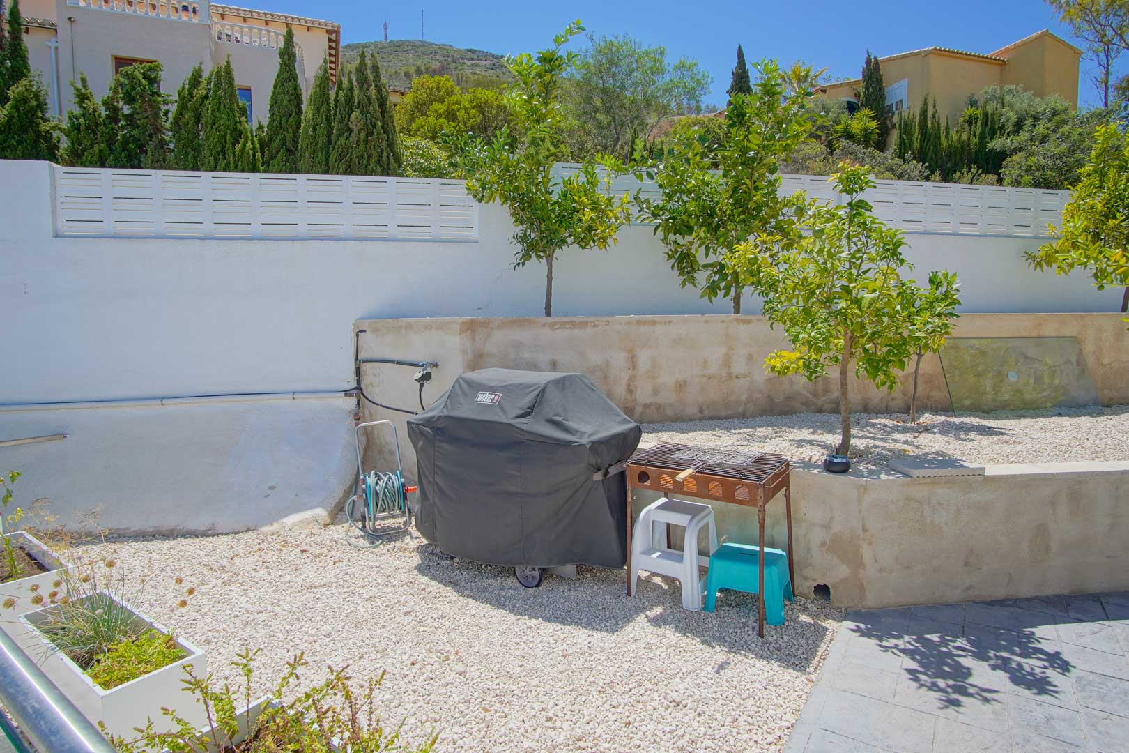 Außenbereich mit weißem Kies, jungen Obstbäumen und Grillstation. Der Garten ist von einer weißen Mauer umgeben und bietet Blick auf die Umgebung.