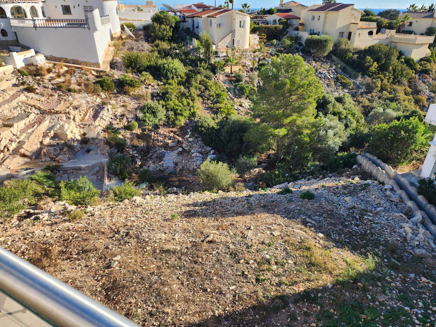 Blick vom Balkon auf das abfallende Grundstück mit Kiesboden, natürlichen Felsformationen und einheimischer mediterraner Bepflanzung.