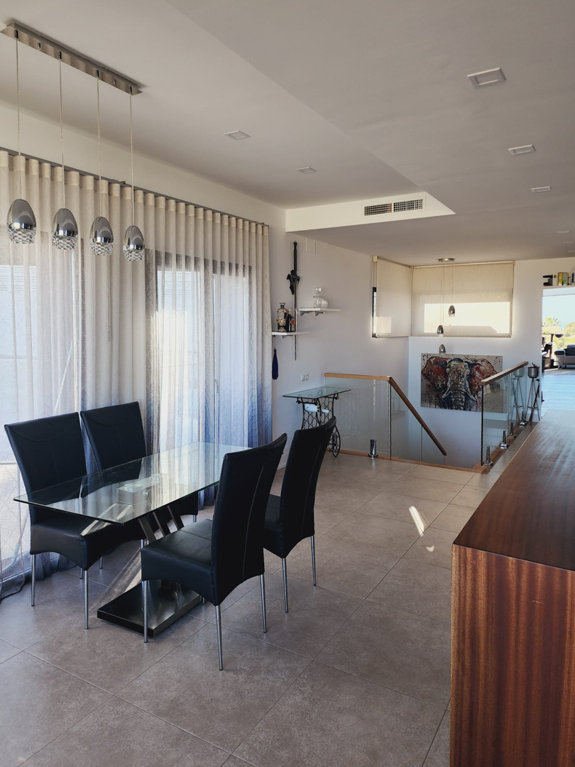 Essbereich mit Glastisch und schwarzen Stühlen neben einer Treppe mit Glasgeländer. Moderne Pendelleuchten und Deckeneinbaustrahler sorgen für ein helles Ambiente.