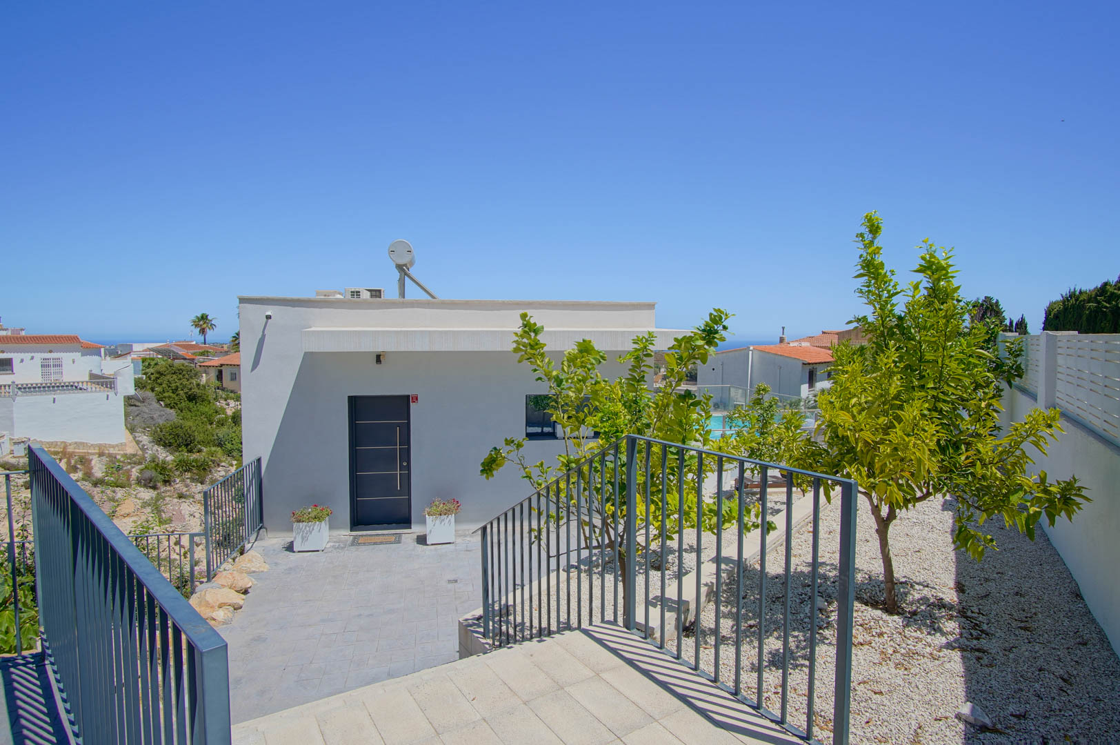 Haupteingang der Villa mit grauer Sicherheitstür, moderner weißer Fassade, Solarthermieanlage und Kiesgarten mit Zitrusbäumen.