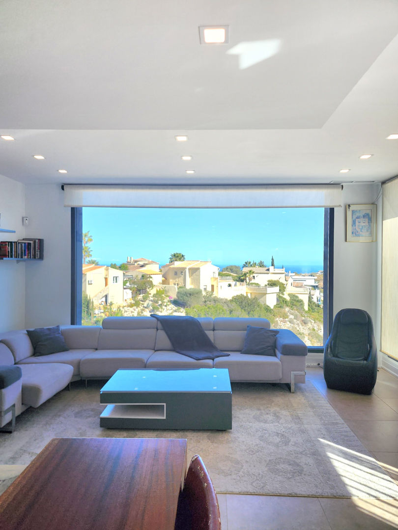 Lichtdurchfluteter Wohnbereich mit großem Panoramafenster zum Meer, einem grauen Ecksofa und moderner LED-Deckenbeleuchtung.