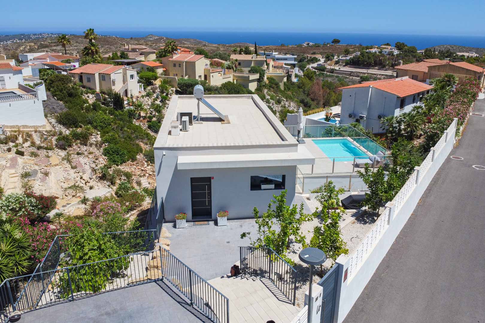 Luftaufnahme der modernen Villa mit Flachdach, Solarthermieanlage und Poolbereich mit Glasgeländer, eingebettet in die Küstenlandschaft mit Meerblick.