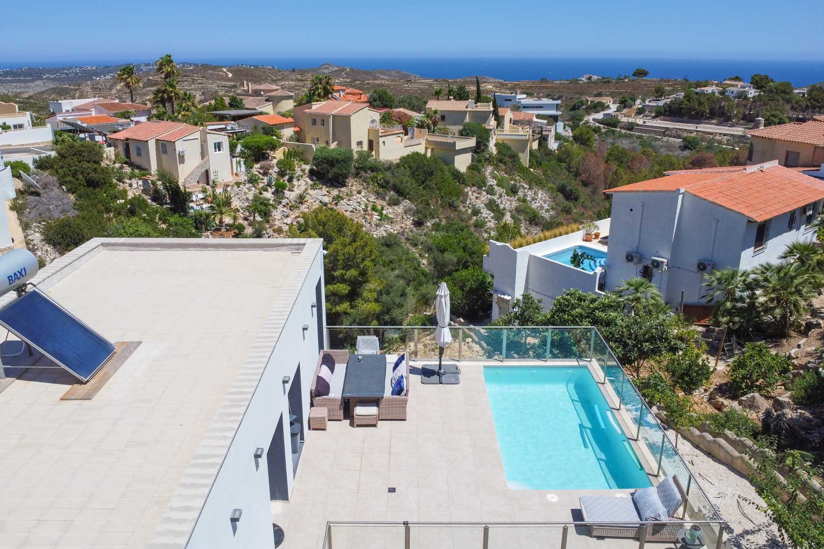 Luftaufnahme der Terrasse mit Pool, Glasgeländern und Außenmöbeln. Das Flachdach umfasst eine Solaranlage und bietet einen weiten Blick auf das Mittelmeer.