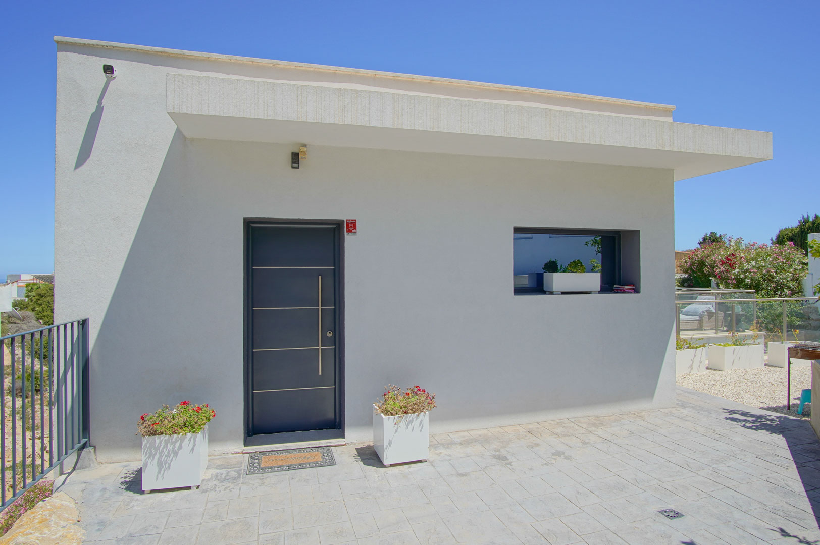 Minimalistische weiße Fassade mit dunkelgrauer moderner Eingangstür, vertikalem Edelstahlgriff und dekorativen Pflanzgefäßen auf dem gepflasterten Zugang.