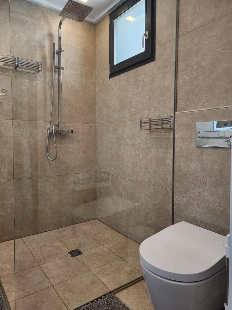 Modernes Badezimmer mit begehbarer Dusche, Glastrennwand, verchromter Regenduscharmatur und sandfarbenen Keramikfliesen an Wänden und Boden.