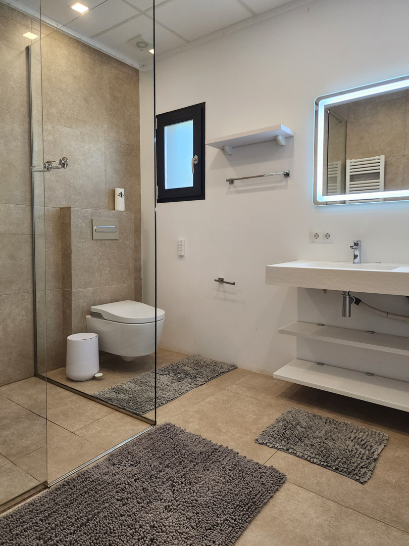 Modernes Badezimmer mit begehbarer Glasdusche, wandhängendem WC und schwebendem Waschtisch mit offenen Regalen sowie einem LED-hintergrundbeleuchteten Spiegel.