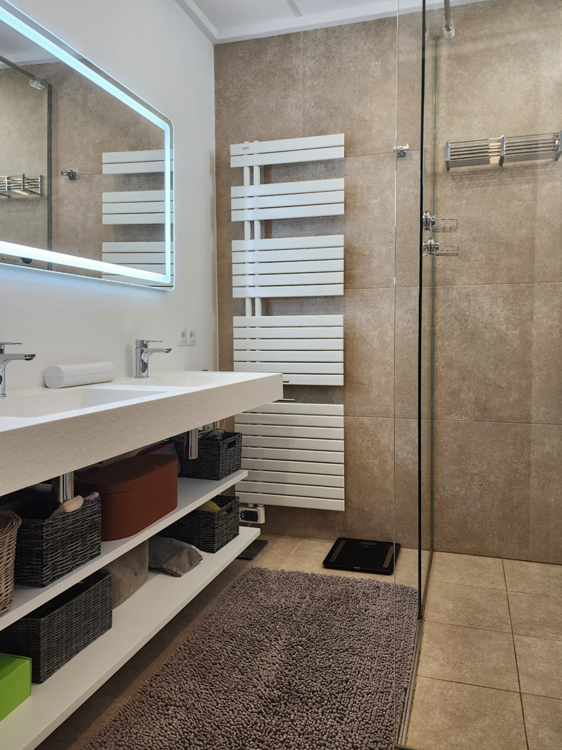 Modernes Badezimmer mit Doppelwaschtisch, offenen Regalen und LED-Spiegel. Ausgestattet mit einem weißen Handtuchheizkörper und einer begehbaren Dusche mit erdigen Fliesen.