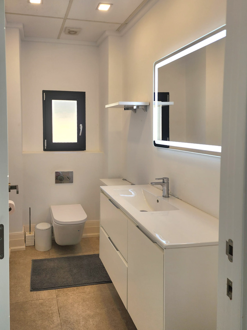 Modernes Badezimmer mit weißem Waschtisch, LED-hinterleuchtetem Spiegel und wandhängendem WC. Es verfügt über Deckeneinbaustrahler und neutrale Bodenfliesen.