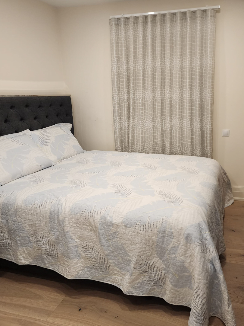 Modernes Schlafzimmer mit Doppelbett, dunkelgrau gestepptem Kopfteil, hellem Holzboden und deckenhohen gemusterten Vorhängen.