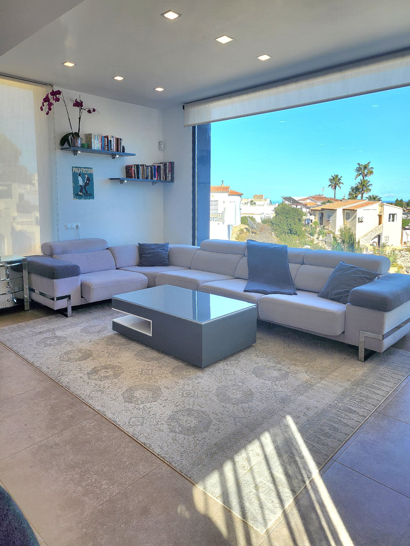 Modernes Wohnzimmer mit L-Sofa, minimalistischem Couchtisch und großem Panoramafenster. Mit Deckeneinbaustrahlern, Schweberegalen und Feinsteinzeugböden.