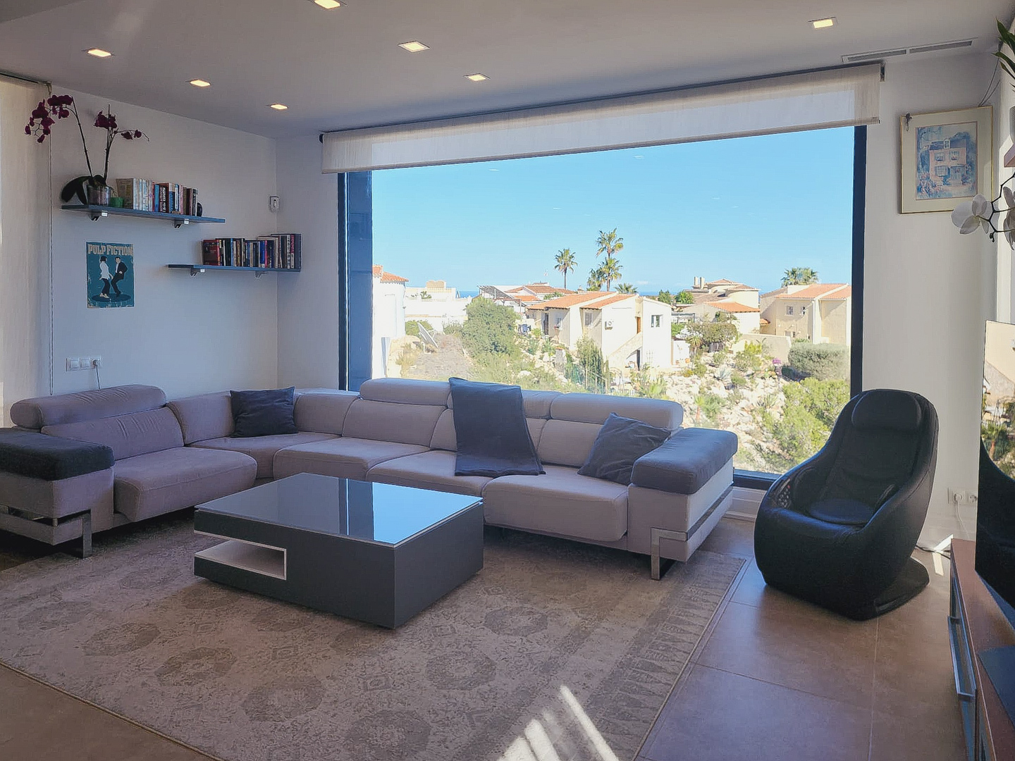 Modernes Wohnzimmer mit L-Sofa, minimalistischem Couchtisch und großem Panoramafenster. Mit Deckeneinbaustrahlern und freiem Blick auf die umliegende Landschaft.
