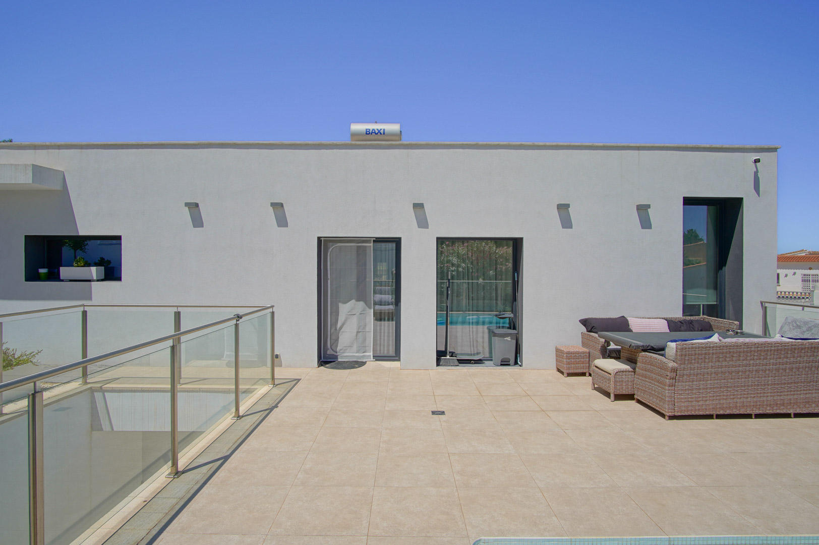 Obere Terrasse mit sandfarbenen Keramikfliesen, Gartenmöbeln aus Polyrattan und Glasgeländern. Auf dem Flachdach ist eine BAXI-Solarthermieanlage installiert.