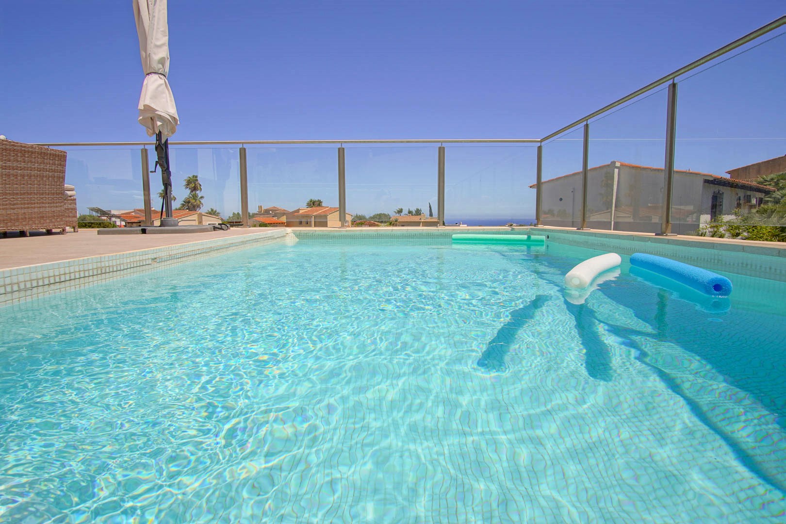 Rechteckiger Pool mit hellblauen Mosaikfliesen und einer Sicherheitsglas-Balustrade. Die Terrasse bietet einen freien Blick auf das Meer und ist mit Gartenmöbeln ausgestattet.