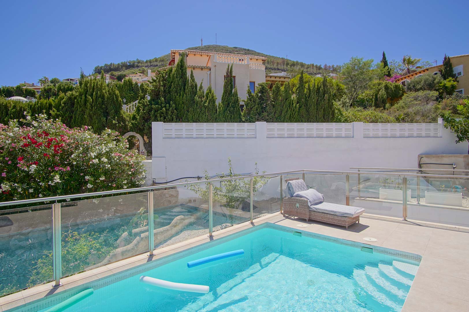 Sonnige Terrasse mit rechteckigem Pool, Glasgeländern und einer Rattanliege, eingebettet in eine Gartenlandschaft mit Oleander und Zypressen.