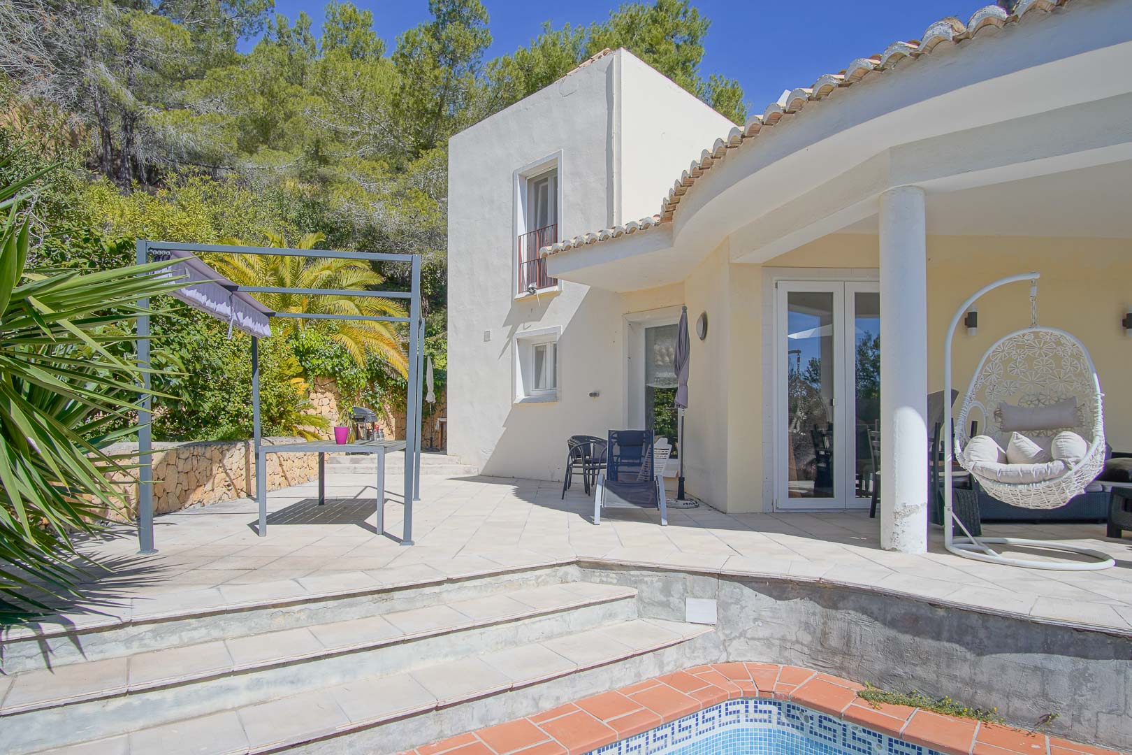 Außenbereich mit Poolrand, Pergola-beschattetem Essplatz und überdachter Veranda mit Hängesessel, umgeben von mediterraner Bepflanzung und Pinien.