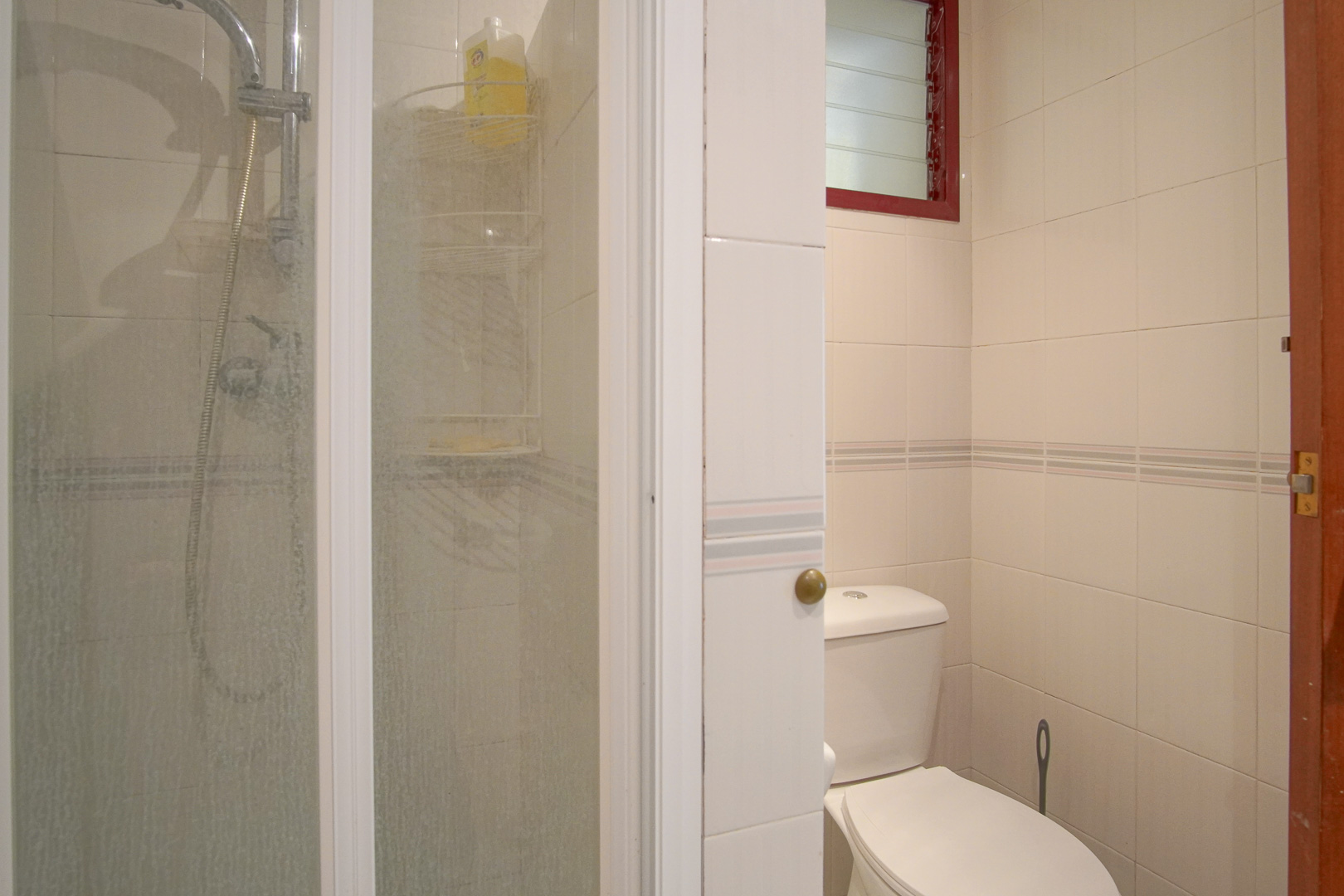 Funktionales Badezimmer mit weiß gefliesten Wänden, einer Glasduschkabine und einem hochgelegenen Lüftungsfenster mit rotem Rahmen.