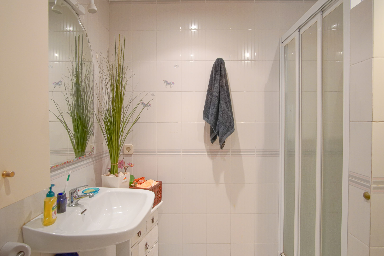 Funktionales Badezimmer mit weiß gefliesten Wänden, Porzellanwaschbecken, Bogenspiegel und Dusche mit weißer Schiebetür-Einfassung.