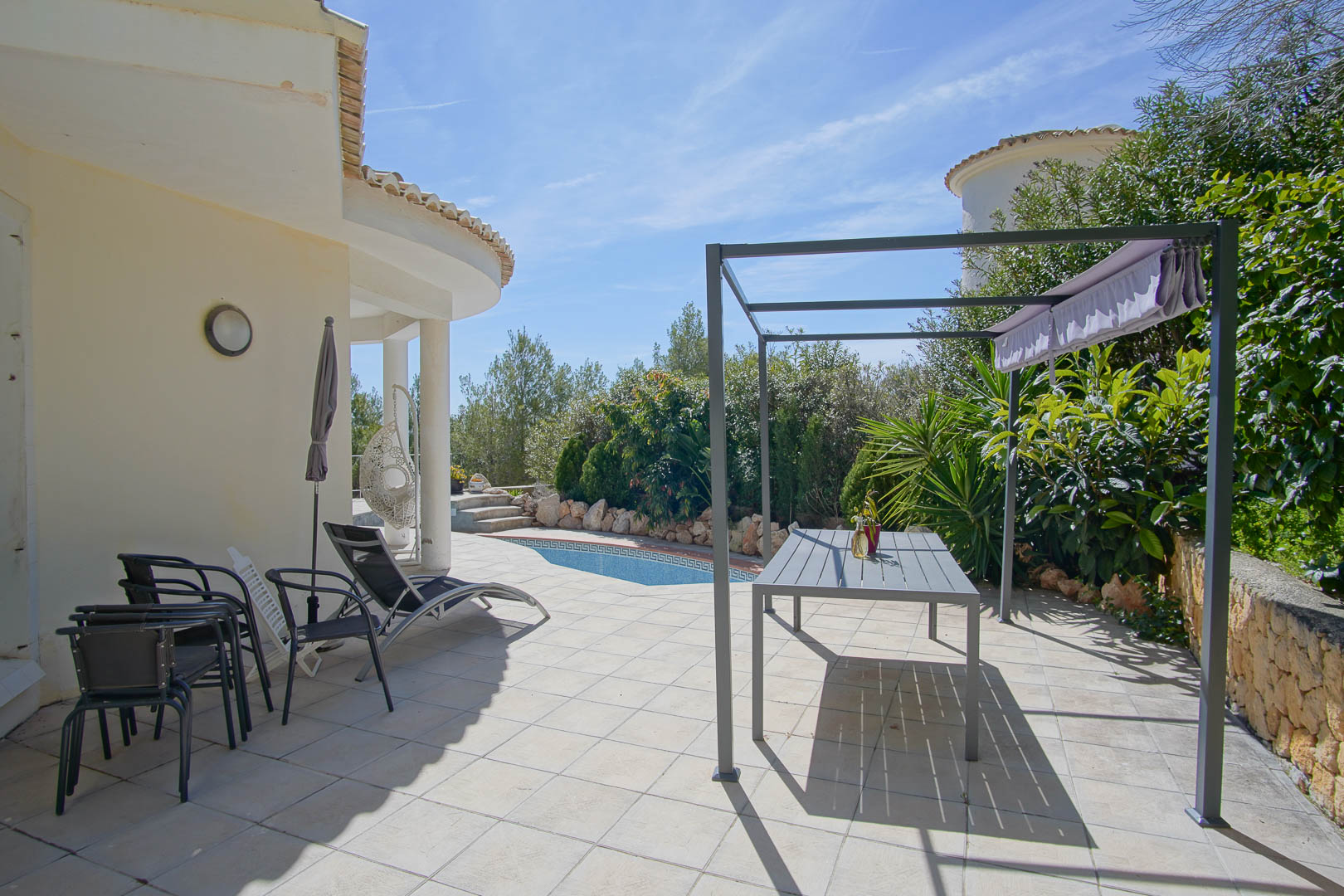 Geflieste Außenterrasse mit Metall-Pergola, Essbereich und privatem Pool, umgeben von gepflegter mediterraner Bepflanzung.