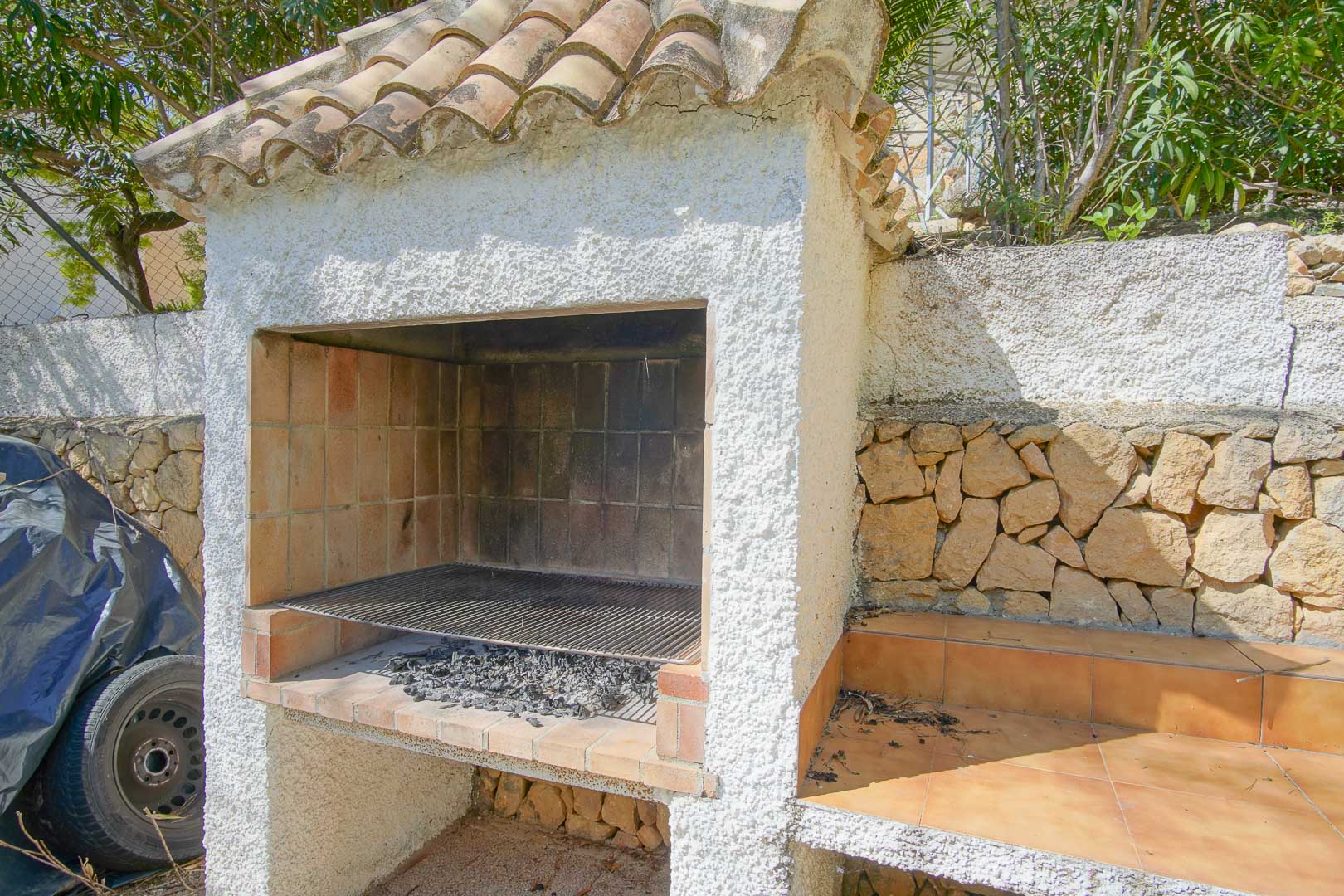 Gemauerter Außengrill mit Terrakotta-Ziegeldach und weißer Putzfassade, integriert an einer Natursteinmauer mit angrenzender gefliester Arbeitsfläche.