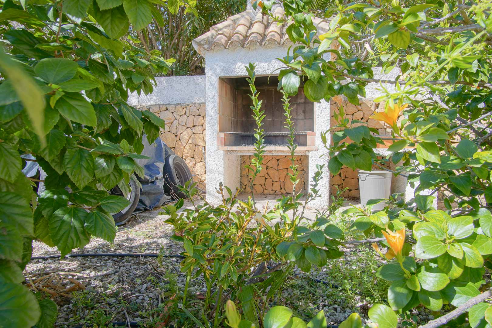 Gemauerter Außengrill mit Ziegeldach, eingebettet in einen mediterranen Garten mit Natursteinmauern und üppiger Bepflanzung.