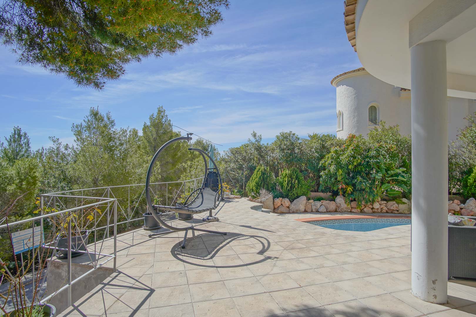 Gepflasterte Terrasse mit modernem Hängesessel, direktem Zugang zum Pool mit Mosaikrand und Blick auf die umliegenden Pinien und Gartenanlagen.