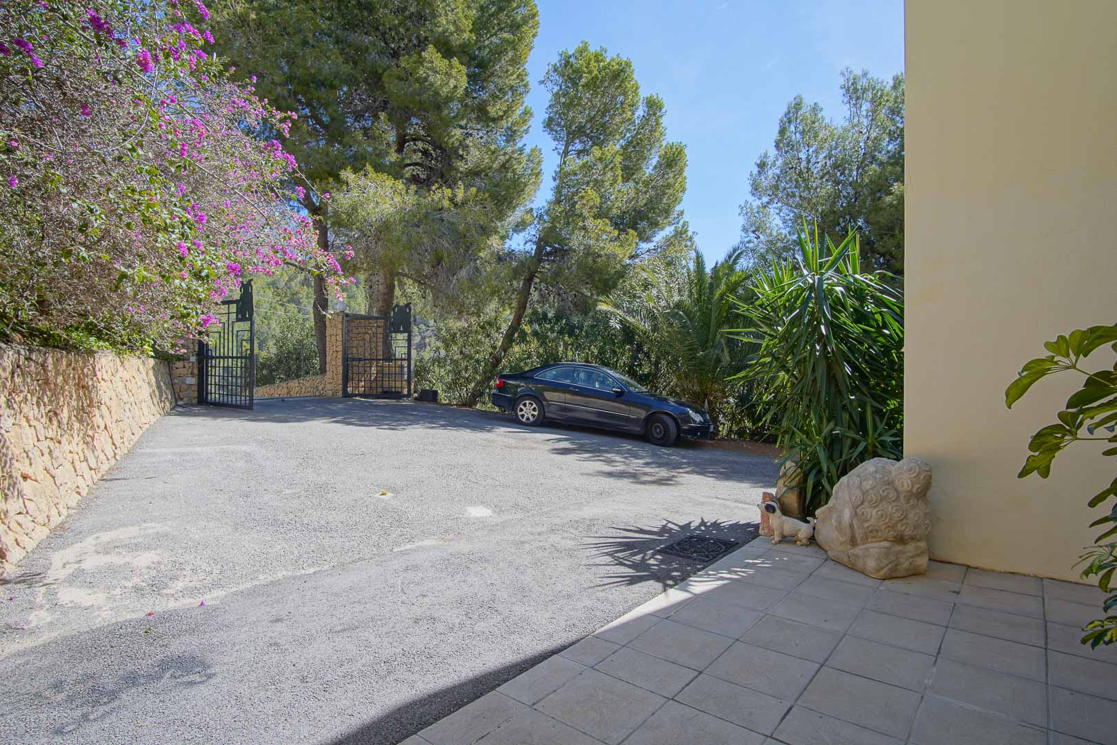 Gesicherte Einfahrt mit Metalltor, großzügigem gepflastertem Parkbereich und Natursteinmauern, umgeben von altem Baumbestand.