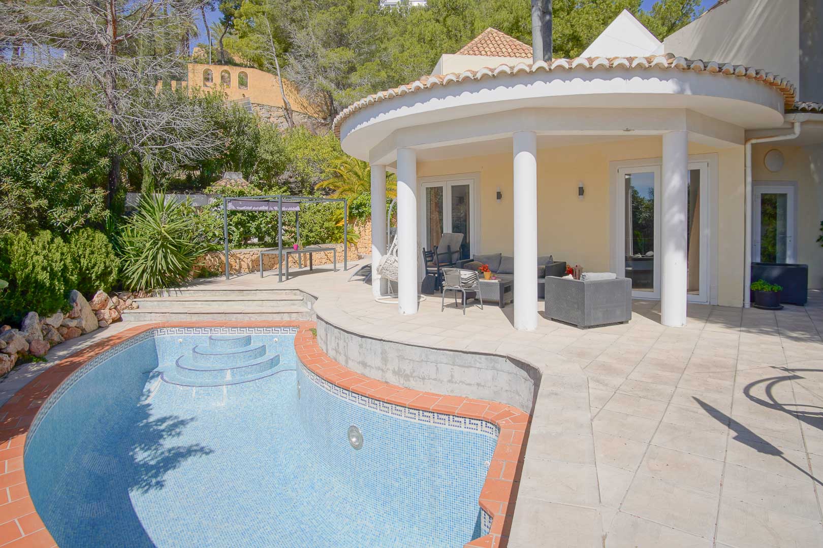 Halbrunde überdachte Terrasse mit weißen Säulen und Blick auf den privaten Pool mit Terrakotta-Einfassung, umgeben von mediterraner Bepflanzung.